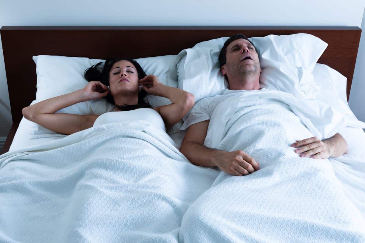 snoring-solutions A heterosexual couple in bed. The man on the right has his mouth open as though snoring. The woman on the left is placing ear plugs into her ears.