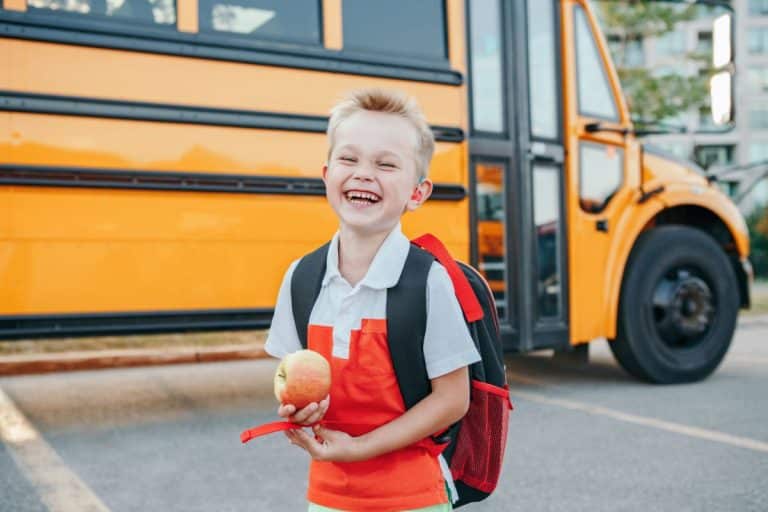 Pediatric Child Hearing Aid Fitting Child with a visible hearing aid standing in front of a school bus. He is holding an apple and has his backpack on. He smiling and mid-laugh.